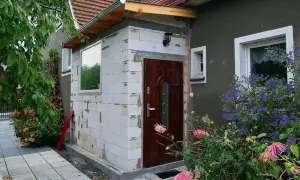 Dobudówka do starego domu w trakcie budowy. Białe pustaki, nowe drzwi i okno czekają na wykończenie. Wokół kwitną kwiaty.