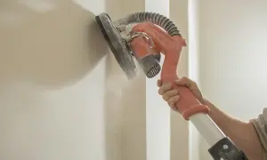 Ręka trzyma żyrafę do gipsu, która szlifuje ścianę. Pył unosi się w powietrzu.