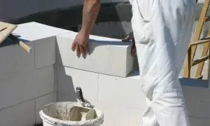 Murarz w białych spodniach układa pustak na budowie. Obok wiadro z zaprawą i kielnia.