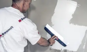Fachowiec w białej czapce i koszulce z logo Baumit wykonuje gipsowanie ścian, używając szerokiej pacy do rozprowadzania masy.