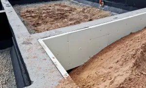Budowa domu w toku. Betonowe stopy fundamentowe otaczają wykop, w którym widać izolację ze styropianu i piasek.