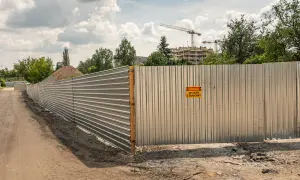 Metalowe ogrodzenie budowy, spełniające wymagania bezpieczeństwa, z tabliczką "Uwaga, wyjazd z budowy". W tle budynki i dźwigi.