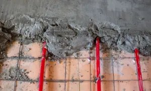Czerwone rury podłogówki ukryte pod mokrym betonem. Trwa praca nad tym, jaka grubość wylewki na podłogówkę będzie optymalna.