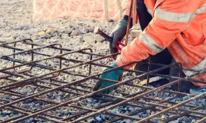 Budowlaniec w pomarańczowej kamizelce zbroi fundament, łącząc pręty. Zbrojenie rozproszone tworzy solidną siatkę.