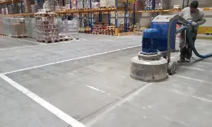 Pracownik wykonuje szlifowanie betonu w magazynie, używając maszyny z odkurzaczem.