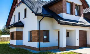 Nowy dom z białym tynkiem i drewnianymi akcentami. Wokół domu widoczne są ciemne cokoły, które dodają mu charakteru.