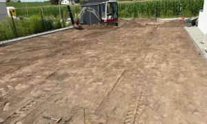 Mini koparka pracuje przy niwelacji terenu pod budowę. W tle widać domy i pole kukurydzy.