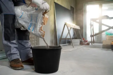 Budowlaniec wsypuje zaprawę do wiadra, przygotowując się do wylewki na styropian. Kluczowe jest dobranie odpowiedniej grubości wylewki, np. ile cm wylewki na styropian.