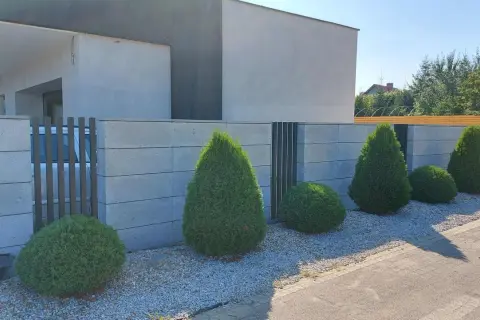 Nowoczesne ogrodzenie murowane tynkowane z betonowych bloczków, otaczające posesję z zadbanym ogrodem i geometrycznymi krzewami.
