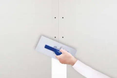 Ręka z kielnią nakłada gips szpachlowy na połączenie płyt gipsowo-kartonowych. Idealny do wygładzania i tworzenia równych powierzchni.