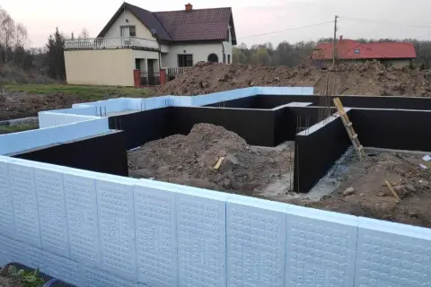 Budowa domu w toku. Widoczna izolacja fundamentów z użyciem płyt XPS. W tle dom jednorodzinny.
