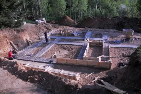 Budowa fundamentów domu. Widać zbrojenie i szalunki. W tle koparka i materiały budowlane. Wylano już warstwę chudego betonu.