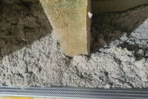 Luźna, szara celuloza wypełnia przestrzeń między drewnianymi belkami, tworząc izolację.