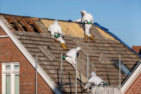 Trzech pracowników w kombinezonach ochronnych usuwa stare pokrycie dachowe, prawdopodobnie zawierające azbest.