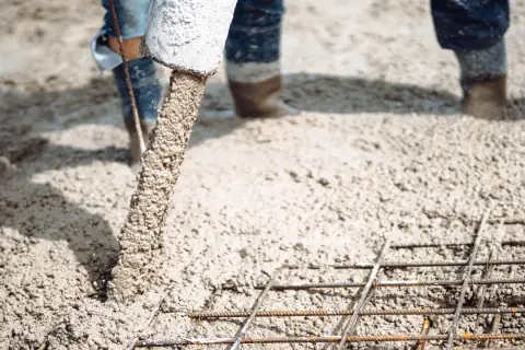 Beton na wylewkę płynie z rury, tworząc solidną podstawę dla budowy. Widać zbrojenie i nogi robotników w gumowcach.