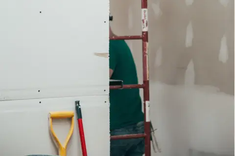 Prace remontowe w toku. Zastanawiasz się, ile schnie gips szybkoschnący? Na pierwszym planie łopata i trzonek, w tle pracownik budowlany.