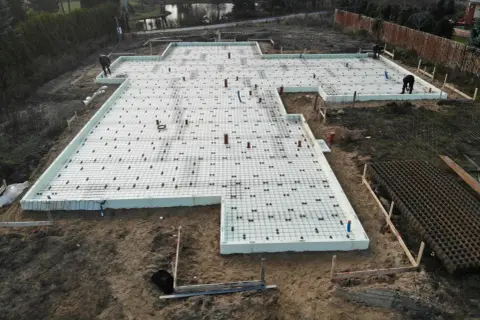 Pracownicy przygotowują zbrojenie płyty fundamentowej. Na białej piance ułożono siatkę stalową, tworząc solidną podstawę pod budowę.