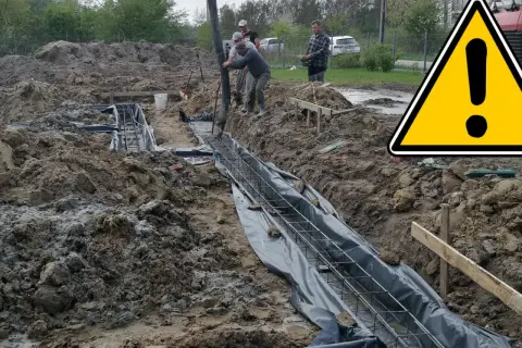 Budowa fundamentów: betonowanie zbrojenia w wykopie, obok żółty znak ostrzegawczy.