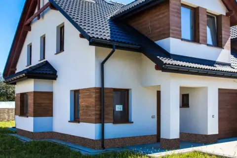 Nowy dom z białym tynkiem i drewnianymi akcentami. Wokół domu widoczne są ciemne cokoły, które dodają mu charakteru.