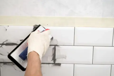 Ręka w rękawiczce nakłada szarą masę na białe płytki, pokazując, czym pomalować fugi.