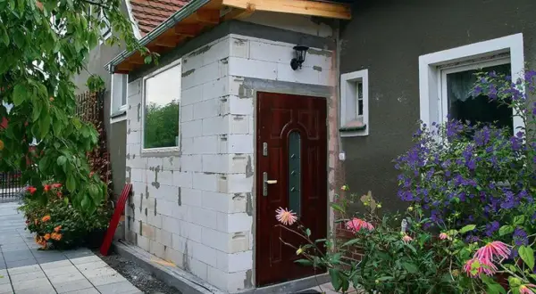 Dobudówka do starego domu w trakcie budowy. Białe pustaki, nowe drzwi i okno czekają na wykończenie. Wokół kwitną kwiaty.