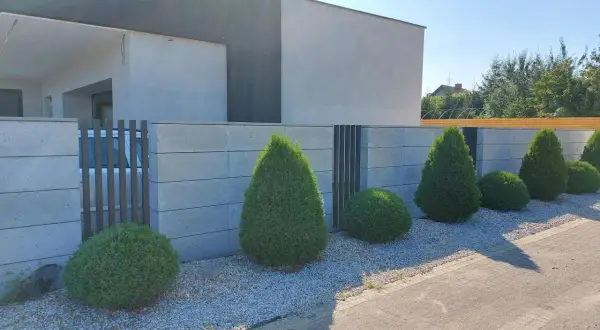 Nowoczesne ogrodzenie murowane tynkowane z betonowych bloczków, otaczające posesję z zadbanym ogrodem i geometrycznymi krzewami.