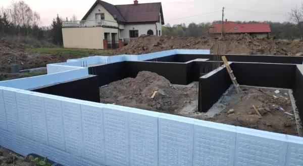 Budowa domu w toku. Widoczna izolacja fundamentów z użyciem płyt XPS. W tle dom jednorodzinny.