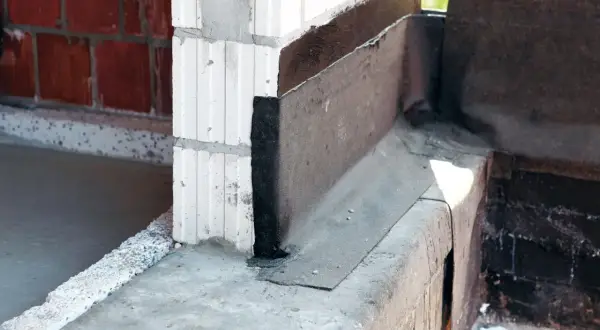 Fragment budowy z widoczną izolacją przeciwwilgociową, czyli papą na fundament. Widać beton, bloczki i cegłę.