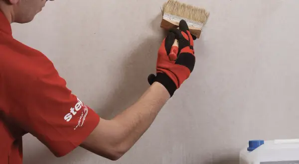 Gruntowanie płyt karton gips to kluczowy etap przygotowania powierzchni. Ręka w rękawicy nakłada preparat pędzlem na ścianę.