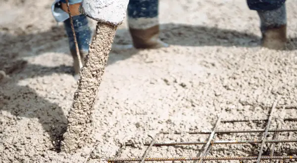 Beton na wylewkę płynie z rury, tworząc solidną podstawę dla budowy. Widać zbrojenie i nogi robotników w gumowcach.