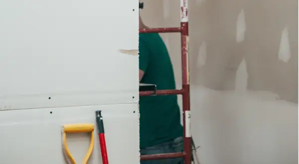 Prace remontowe w toku. Zastanawiasz się, ile schnie gips szybkoschnący? Na pierwszym planie łopata i trzonek, w tle pracownik budowlany.