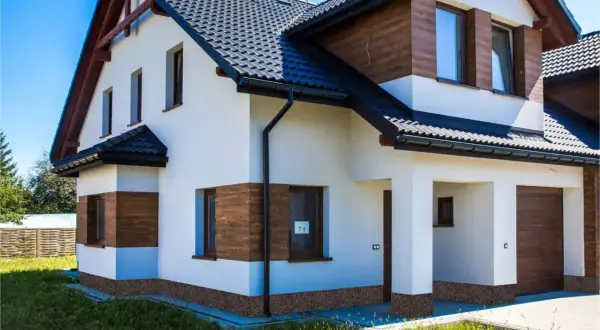 Nowy dom z białym tynkiem i drewnianymi akcentami. Wokół domu widoczne są ciemne cokoły, które dodają mu charakteru.