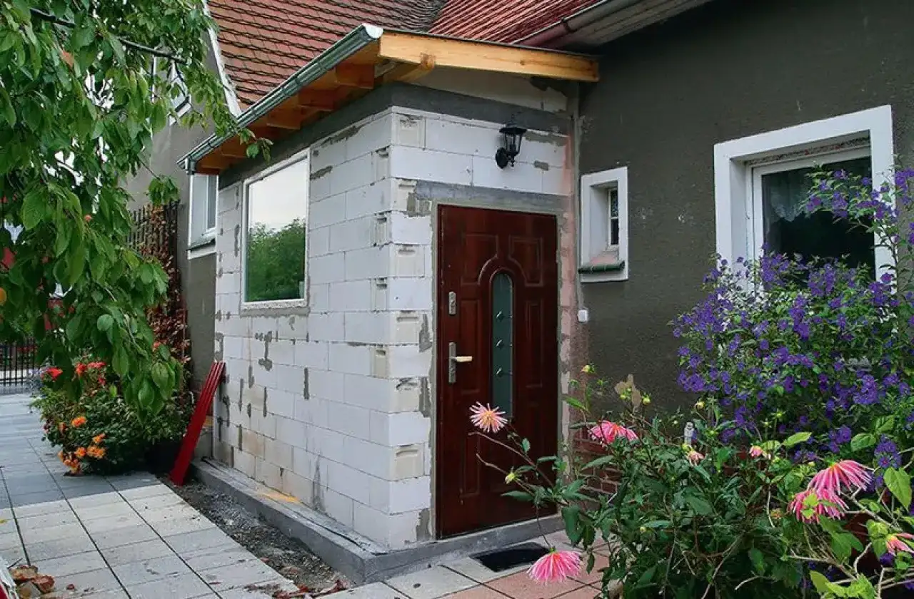Dobudówka do starego domu w trakcie budowy. Białe pustaki, nowe drzwi i okno czekają na wykończenie. Wokół kwitną kwiaty.