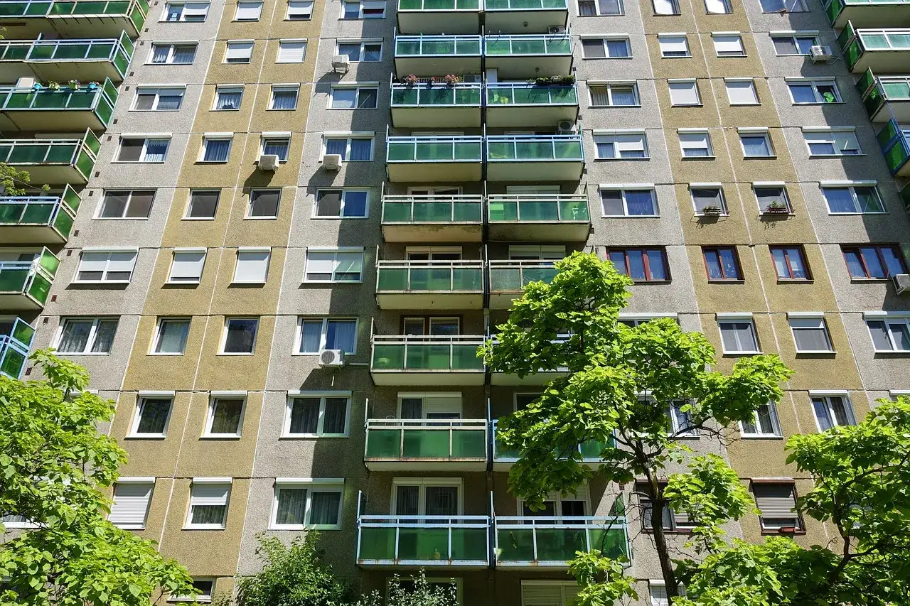 Wielopiętrowe bloki z wielkiej płyty z balkonami i oknami, otoczone zielenią drzew.