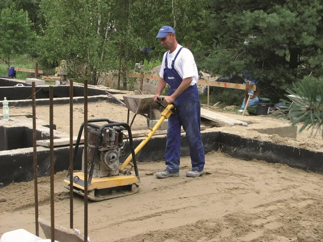 Budowlaniec zagęszcza piasek ubijakiem. W tle widać fundamenty, zbrojenia i materiały budowlane, przygotowane do wylania suchego betonu.