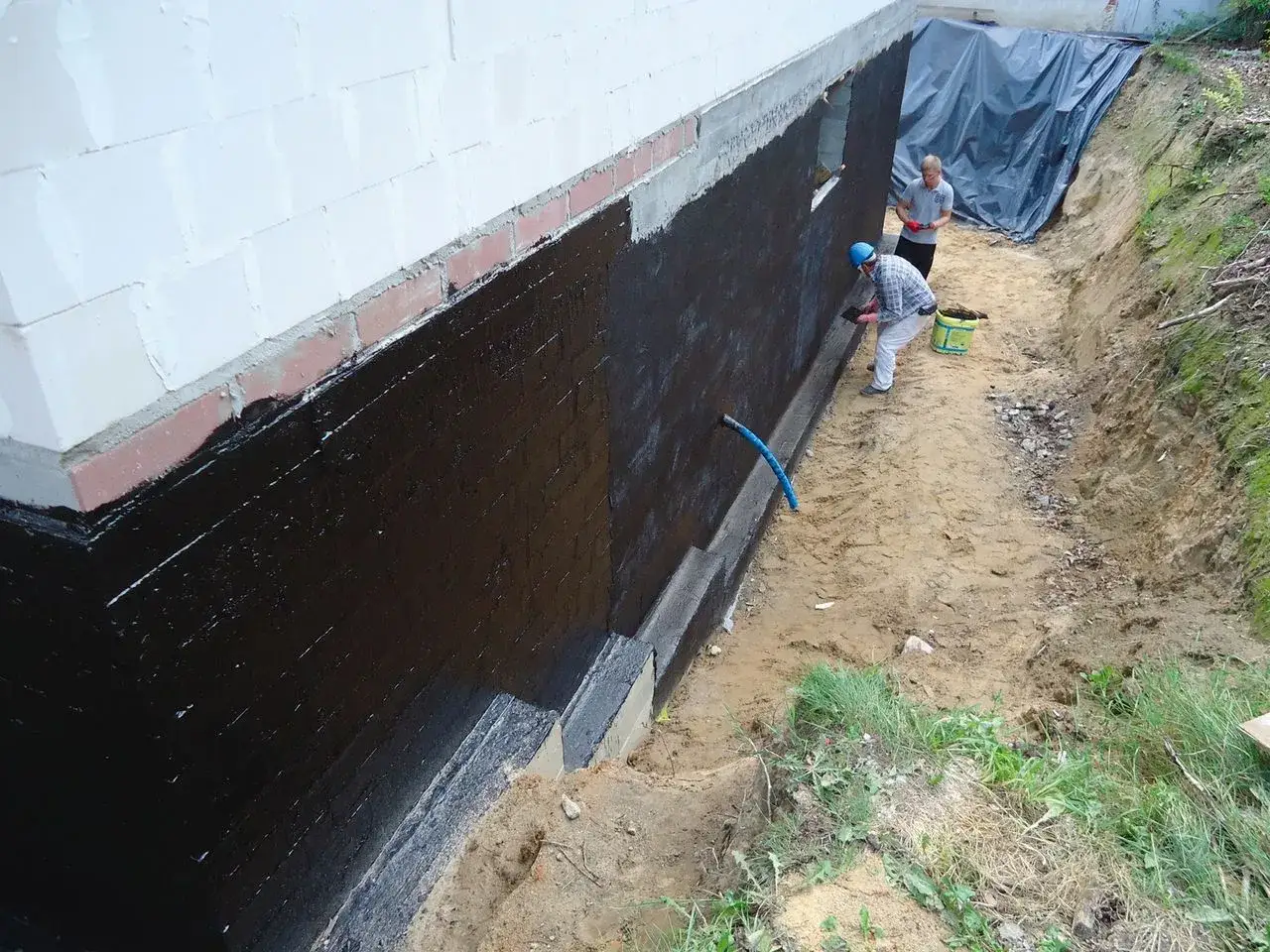 Pracownicy zabezpieczają fundamenty przed wilgocią, nakładając grubą warstwę czarnej masy izolacyjnej. To kluczowy etap, by zapobiec problemom z wilgocią i zapewnić trwałość budowli.