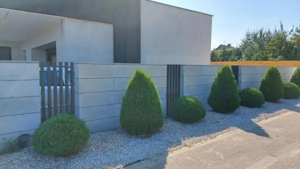 Nowoczesne ogrodzenie murowane tynkowane z betonowych bloczków, otaczające posesję z zadbanym ogrodem i geometrycznymi krzewami.