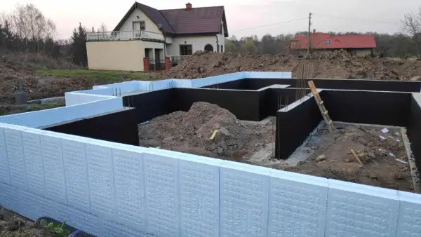 Budowa domu w toku. Widoczna izolacja fundamentów z użyciem płyt XPS. W tle dom jednorodzinny.