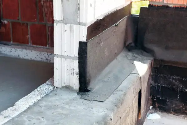 Fragment budowy z widoczną izolacją przeciwwilgociową, czyli papą na fundament. Widać beton, bloczki i cegłę.