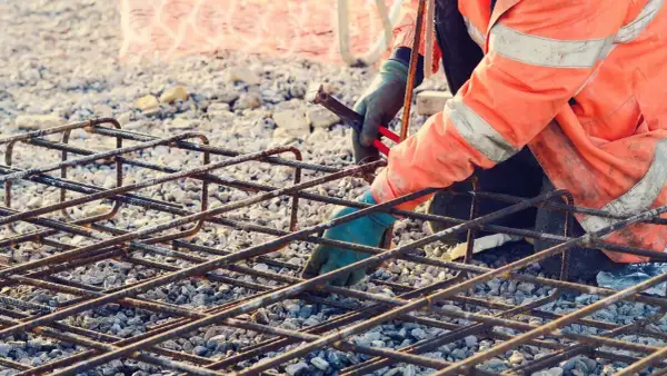 Budowlaniec w pomarańczowej kamizelce zbroi fundament, łącząc pręty. Zbrojenie rozproszone tworzy solidną siatkę.