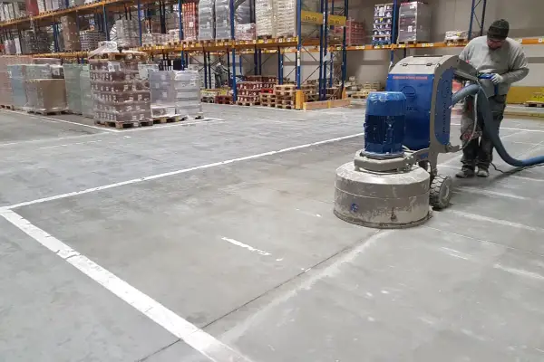 Pracownik wykonuje szlifowanie betonu w magazynie, używając maszyny z odkurzaczem.