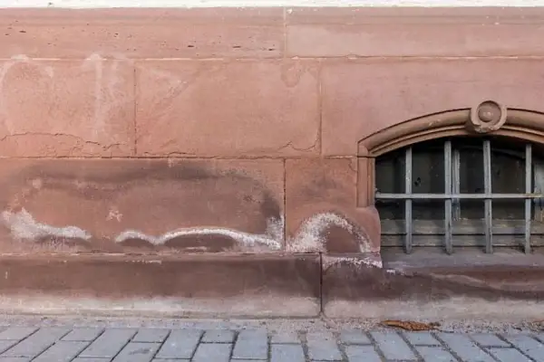 Wilgotne plamy na kamiennym murze piwnicy, wskazujące na potrzebę dowiedzenia się, jak osuszyć fundamenty. Widoczne kraty w małym oknie.