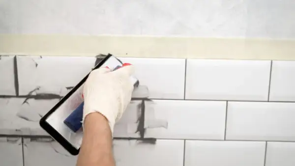 Ręka w rękawiczce nakłada szarą masę na białe płytki, pokazując, czym pomalować fugi.
